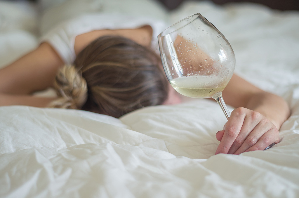 Een vrouw houdt nog net een glas witte wijn vast terwijl ze op haar buik in slaap lijkt te vallen.